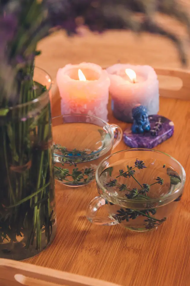 Purple lilacs in a glass vase. Two purple candles. A blue figurine on a purple stone. 2 glasses filled with water and lilac buds. 